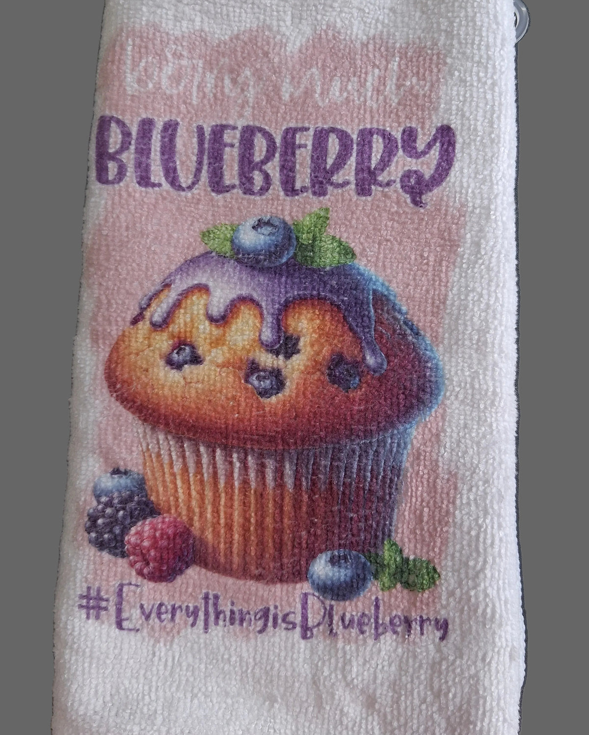 A white textured towel featuring a colorful printed design of a blueberry muffin with the text 'lovin' mail BLUEBERRY' and '#EverythingisBlueberry' on a dark gray background.